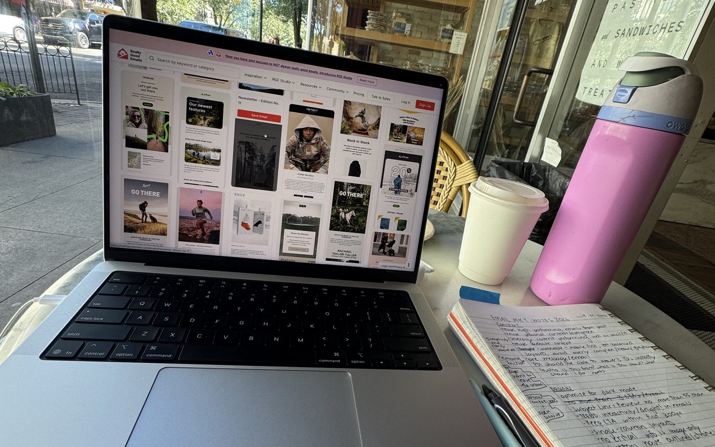 A laptop showing email newsletter designs on a café table next to a coffee cup and handwritten notes